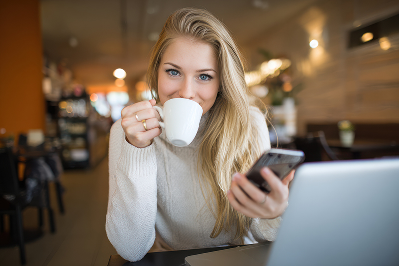 Stock photo of a young woman drinking coffee while working on a laptop and smartphone, with the AI-generated title and 40 keywords Deskria wrote for it